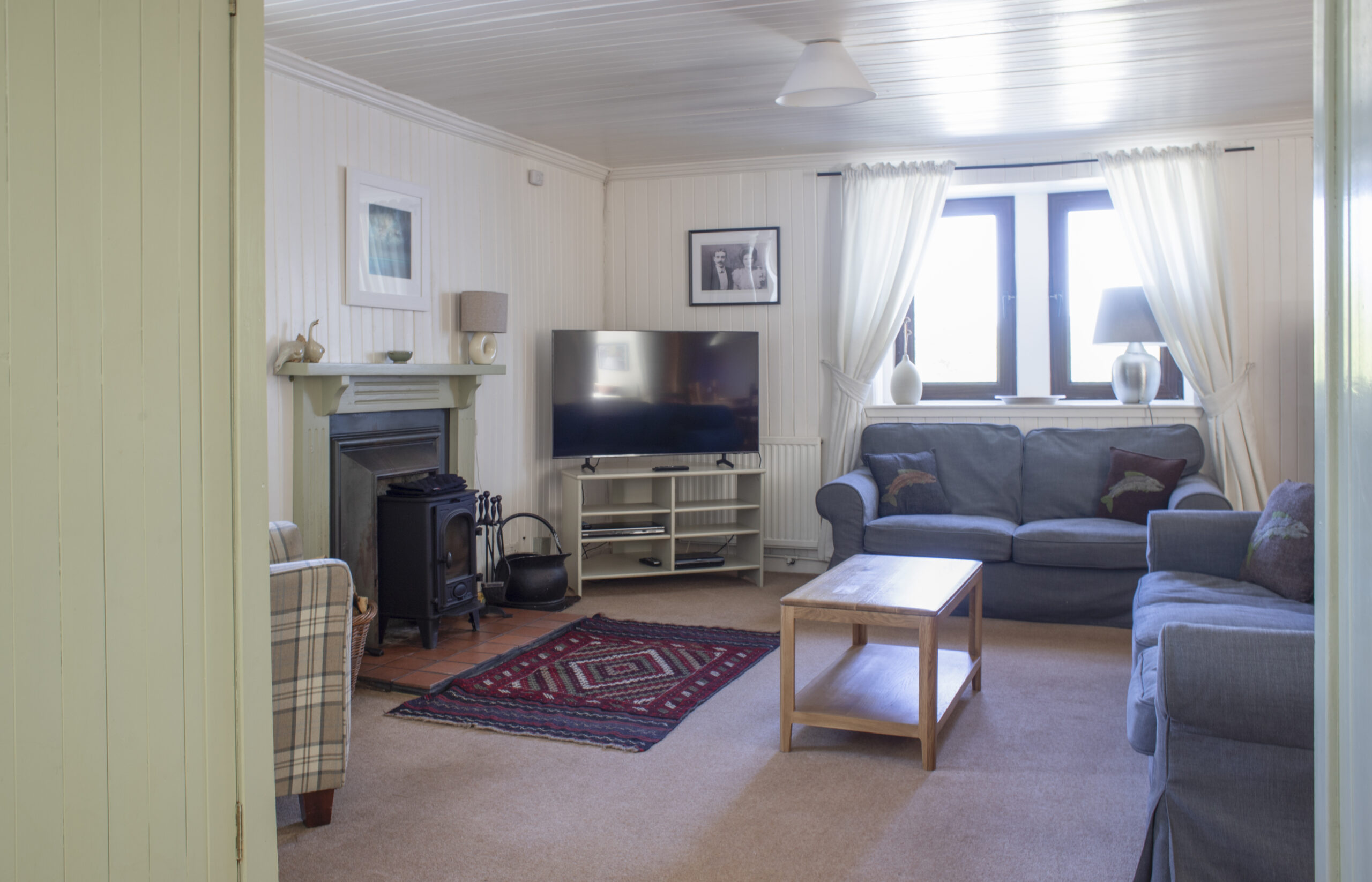 Ploughmans Cottage living room with sofas, carpeted floor, wood burner, large window, TV, coffee table, and arm chair.